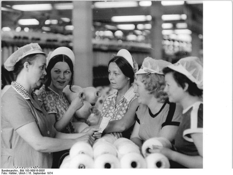 Five women in headscarves at a textile mill, one showing another a spool of thread, Ebersbach, 1974